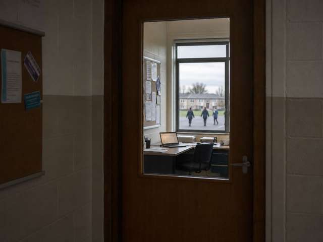 View through a half-glazed staffroom door into a school office: a desk with an open laptop, a noticeboard with printed sheets, and through the far window, the silhouettes of children in a school yard.