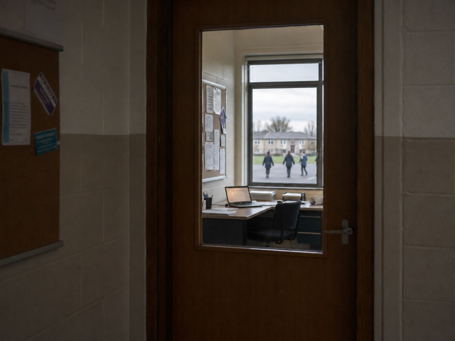 View through a half-glazed staffroom door into a school office: a desk with an open laptop, a noticeboard with printed sheets, and through the far window, the silhouettes of children in a school yard.